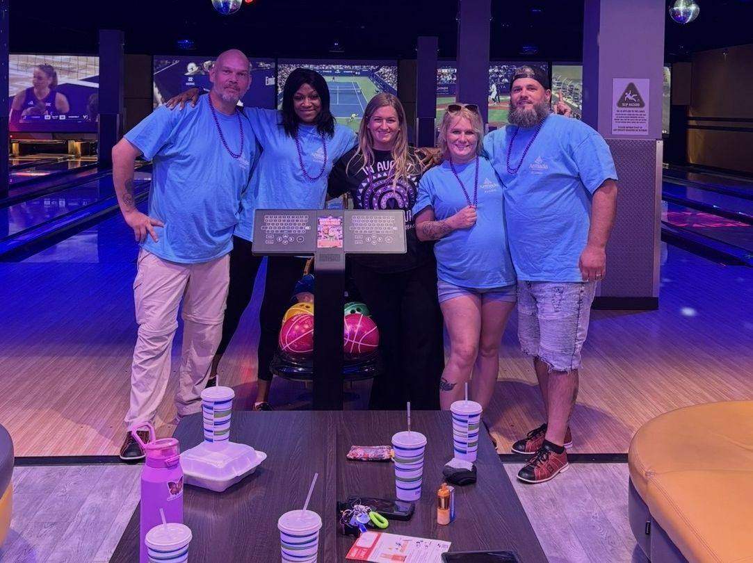 Five people in matching blue shirts with their arms around each other at a bowling alley