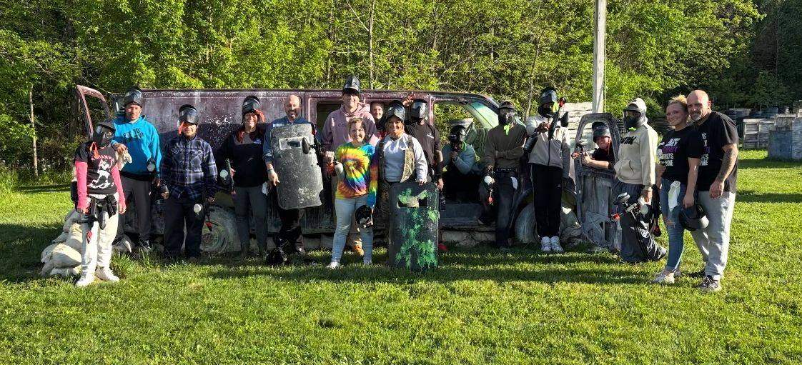 A large group in paintball masks gathered around an old van on a grass field
