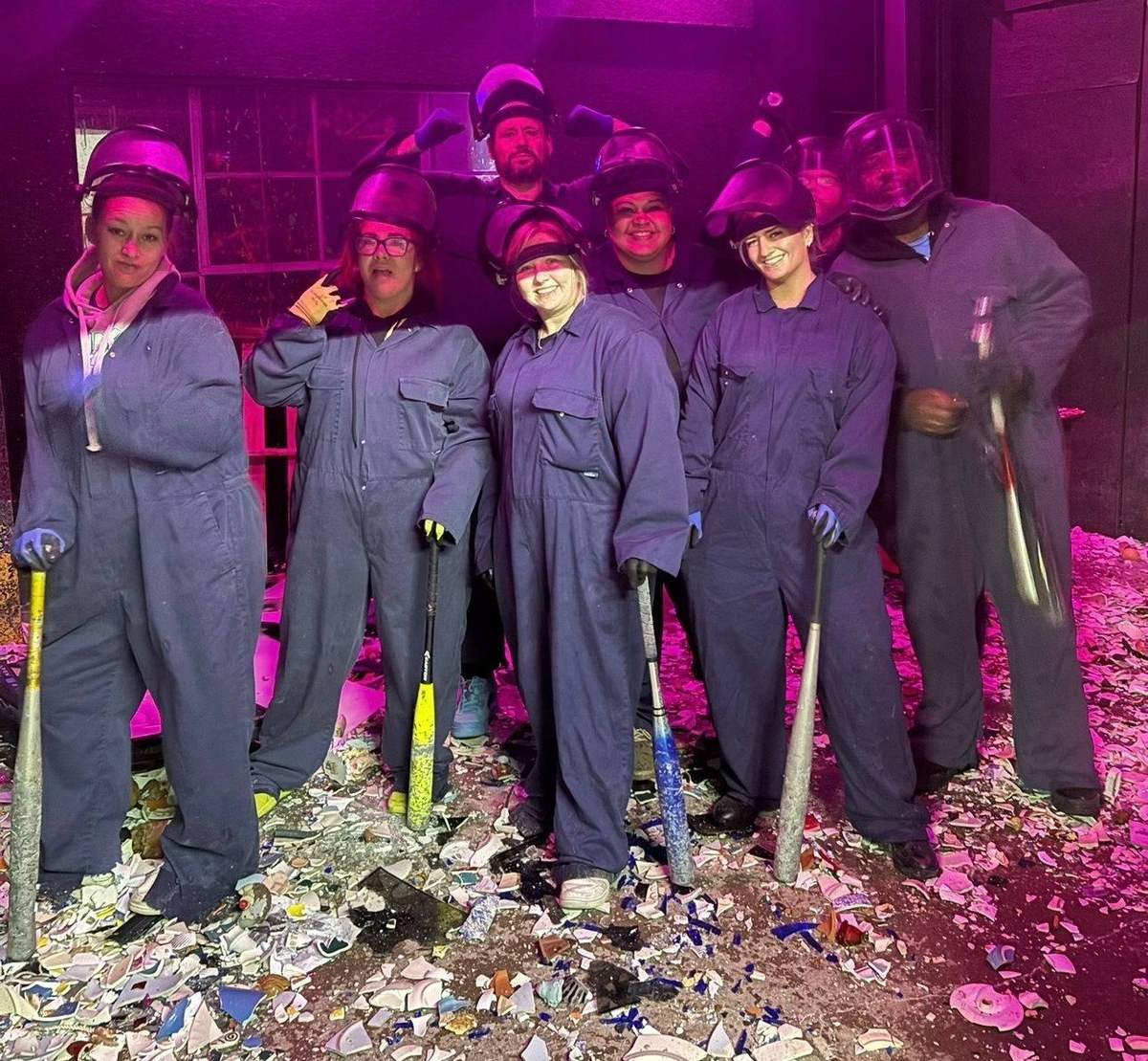 A group in coveralls and face shields holding bats in a rage room, surrounded by broken plates
